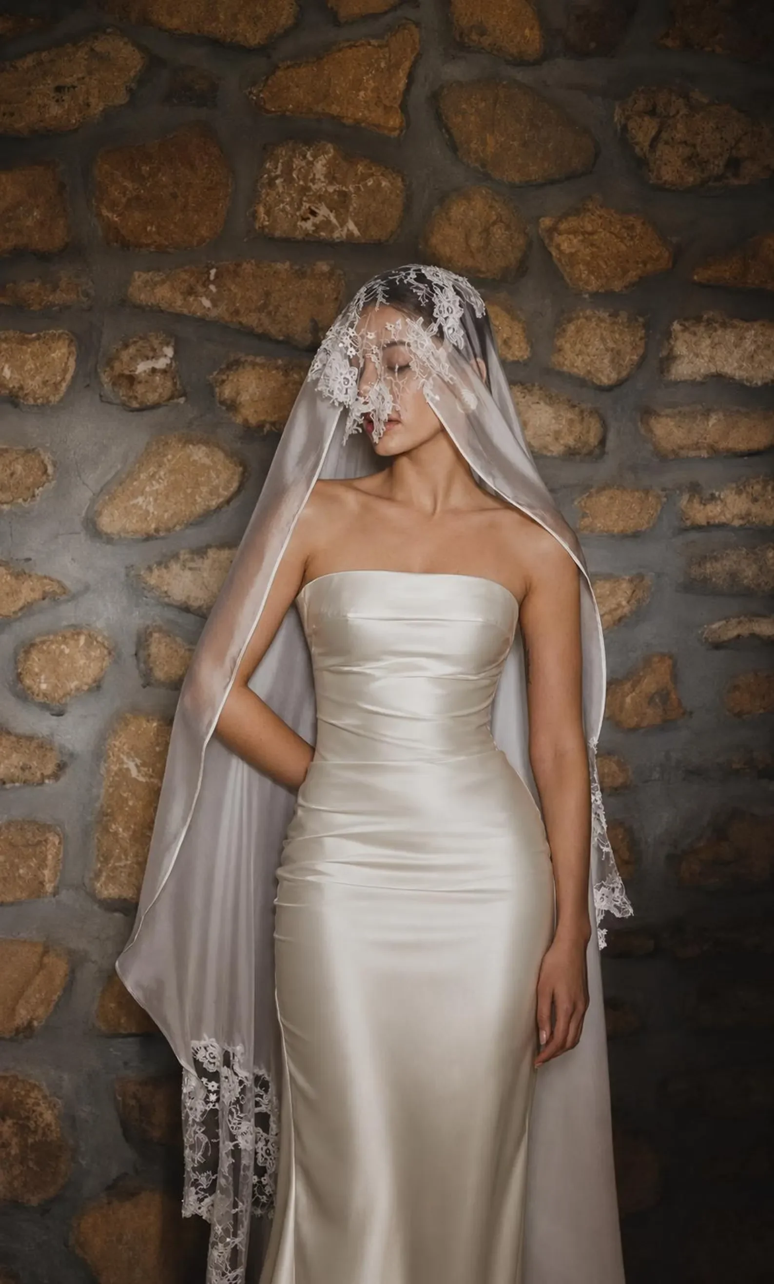 Braut in einem eng anliegenden, trägerlosen Satin-Kleid vor einer Natursteinwand. Ein zarter Schleier mit aufwendigen Spitzen-Applikationen bedeckt schützend ihr Gesicht und ihre Schultern.