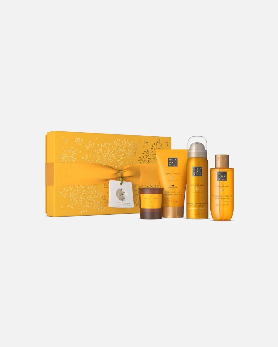 Orangefarbenes Rituals-Geschenkset "The Ritual of Mehr". Vor einer verzierten Box mit Schleife stehen eine kleine braune Kerze und drei Pflegeprodukte.
