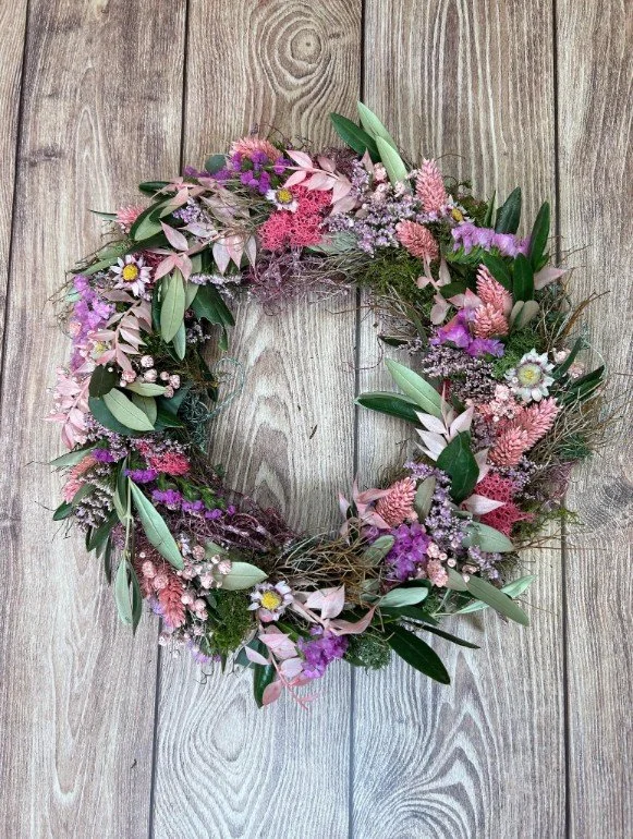 Runder, flach liegender Kranz auf hellbraunem Holzboden. Locker gebunden aus rosé- und lachsfarbenen Trockengräsern, lila Statice, kleinen weiß-pinken Strohblumen und länglichen grünen Olivenblättern auf moosiger Basis.