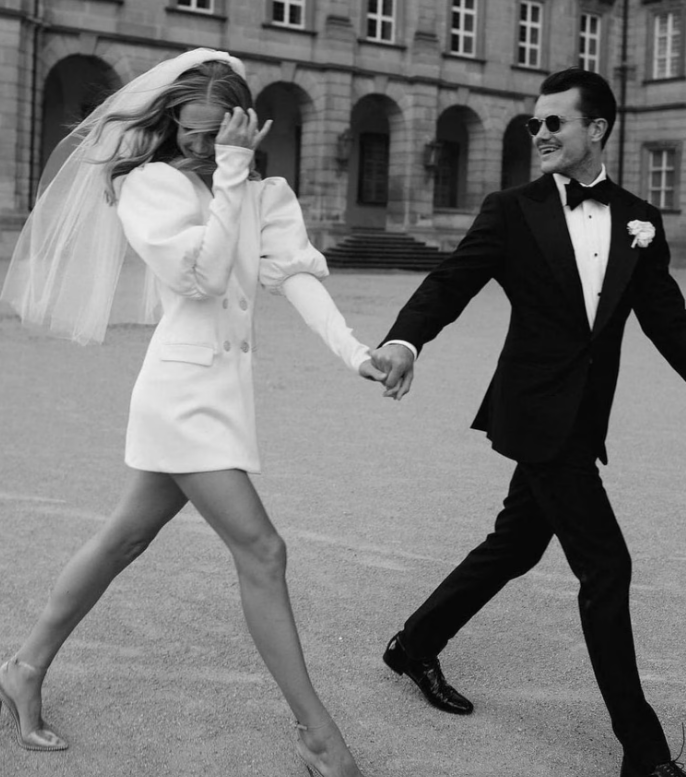 Ein Brautpaar läuft fröhlich Hand in Hand im Freien; die Braut trägt ein kurzes, weißes Kleid mit Schleier, der Bräutigam einen Smoking und Sonnenbrille. Schwarz-Weiß-Bild.
