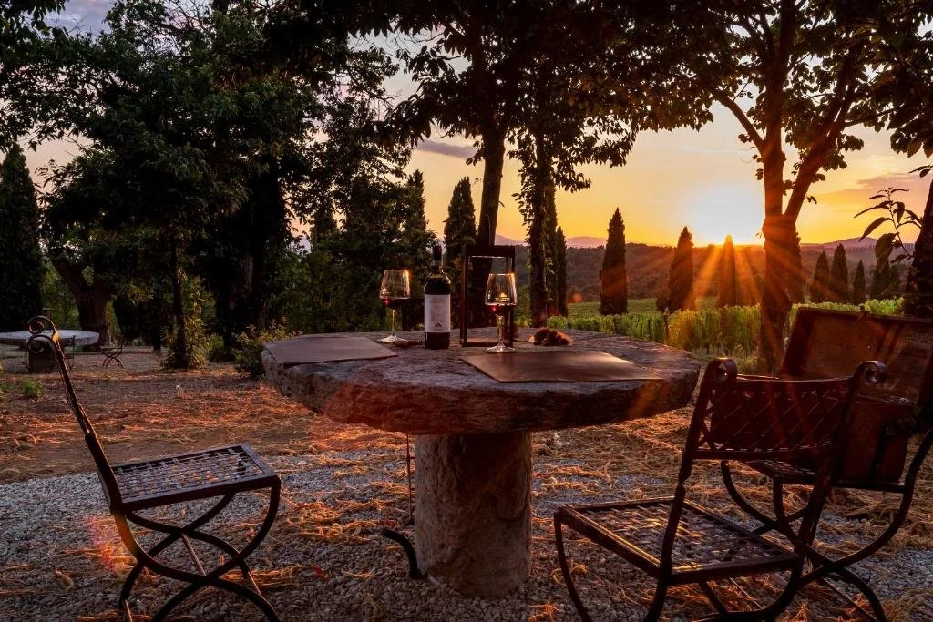 Foto eines rustikalen runden Steintischs mit zwei Gläsern Rotwein im Schatten alter Bäume. Im Hintergrund leuchtet die tiefe Abendsonne über einer toskanischen Landschaft mit Weinbergen und Zypressen.