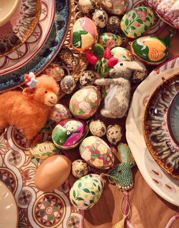 Detailaufnahme bunter Osterdekoration auf dem Tisch: Handbemalte Eier mit Blumen- und Vogelmotiven, Wachteleier sowie kleine Filzfiguren (Hase und Lamm) und ein Perlen-Anhänger.