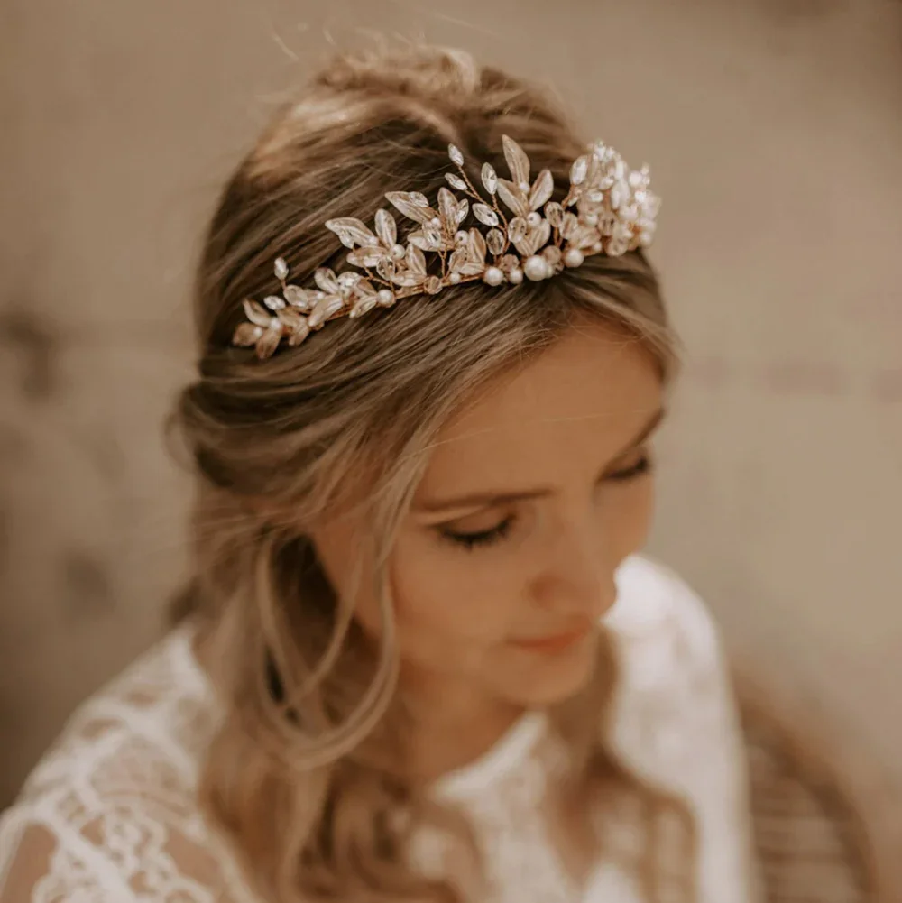 Blonde Braut mit offener, gewellter Frisur und einem goldenen, blätterförmigen Diadem für die Hochzeit in einer romantischen Pose.