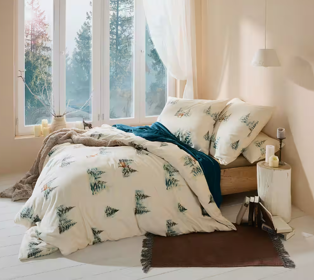 Gemütliches Schlafzimmer mit großem Fenster. Bett mit cremefarbener Bettwäsche mit Tannen- und Hirschmotiven, dunkler Decke, Kissen und einer Plüschdecke auf weißem Dielenboden.