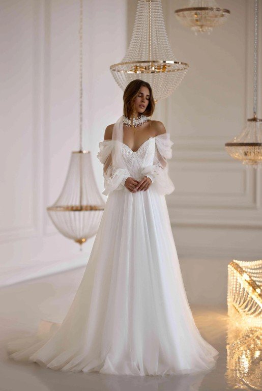 Frau in einem schulterfreien weißen Brautkleid mit zarten Tüllärmeln, stehend in einem eleganten Raum mit Kristallkronleuchtern.