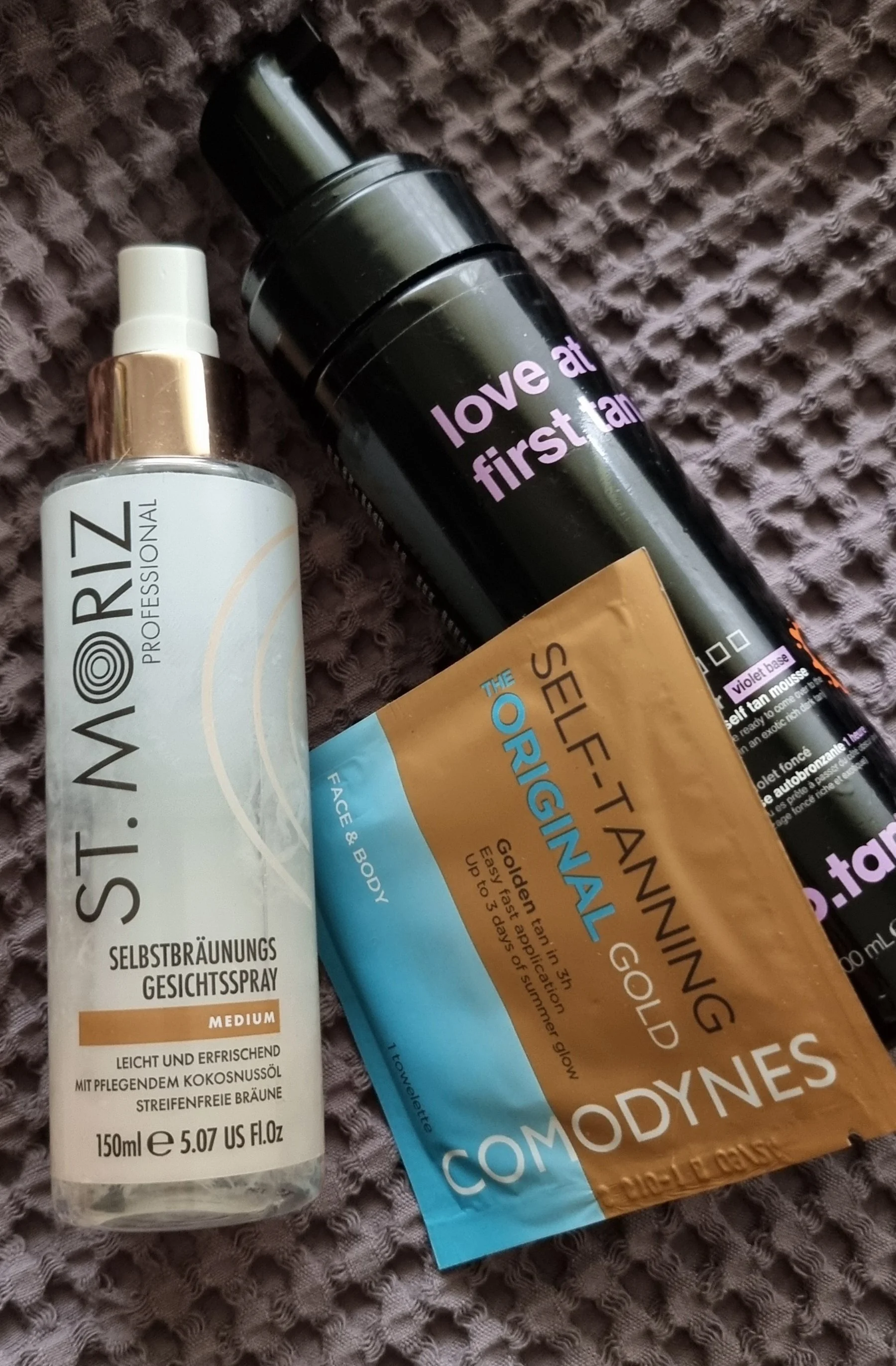 Drei verschiedene Selbstbräuner-Produkte (St. Moriz Gesichtsspray, Comodynes Bräunungstuch und b.tan Mousse) liegen zusammen auf einem grauen Handtuch.