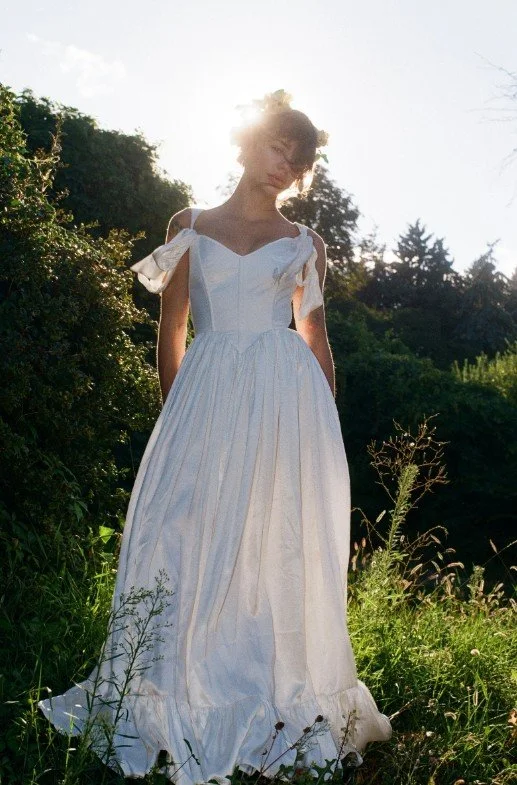 Eine Braut in einem langen, weißen Korsett-Kleid mit weiten, herabhängenden Ärmelschleifen und einem Blumenkranz im Haar steht bei tiefstehender Sonne im hohen Gras. Das starke Gegenlicht erzeugt eine romantische, ländliche Boho-Stimmung.