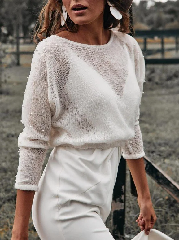 Frau in einem weißen, leicht durchsichtigen Brautpullover mit Perlenbesatz an den Ärmeln, kombiniert mit einer weißen Hose im Freien.