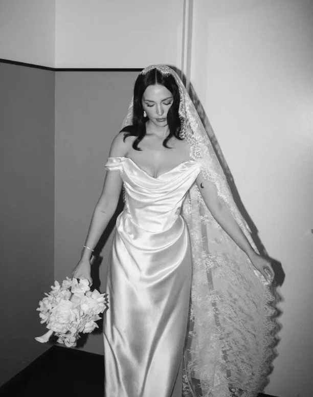 Elegante Braut in einem fließenden, schulterfreien Satin-Brautkleid mit Korsagen-Drapierung und einem traditionellen, langen Mantilla-Schleier aus italienischer Spitze. Inspiration für edle Hochzeitslooks im Holistic Mag.