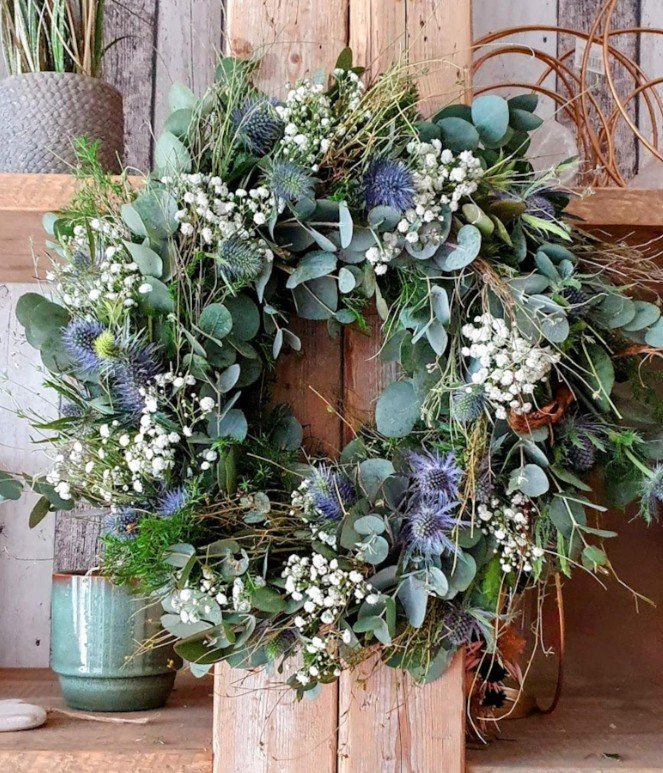 Großer, wild gebundener Blumenkranz an rustikalem Holzbalken: silbrig-grüner Eukalyptus, weißes Schleierkraut und blaue Disteln. Verschwommener Hintergrund mit Floristik und Dekor.