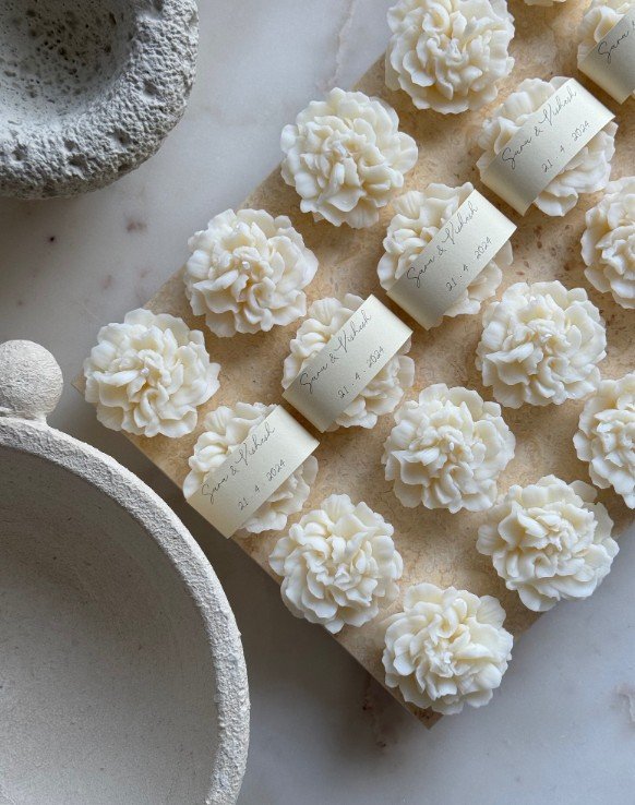 Weiße, blumenförmige Duftkerzen als elegante Gastgeschenke, arrangiert auf einer Steinplatte mit personalisierten Namensetiketten.