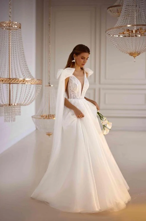 Frau in einem weißen Brautkleid mit tiefem V-Ausschnitt und Tüllrock, mit Blumenstrauß in einem eleganten Raum mit Kronleuchtern.