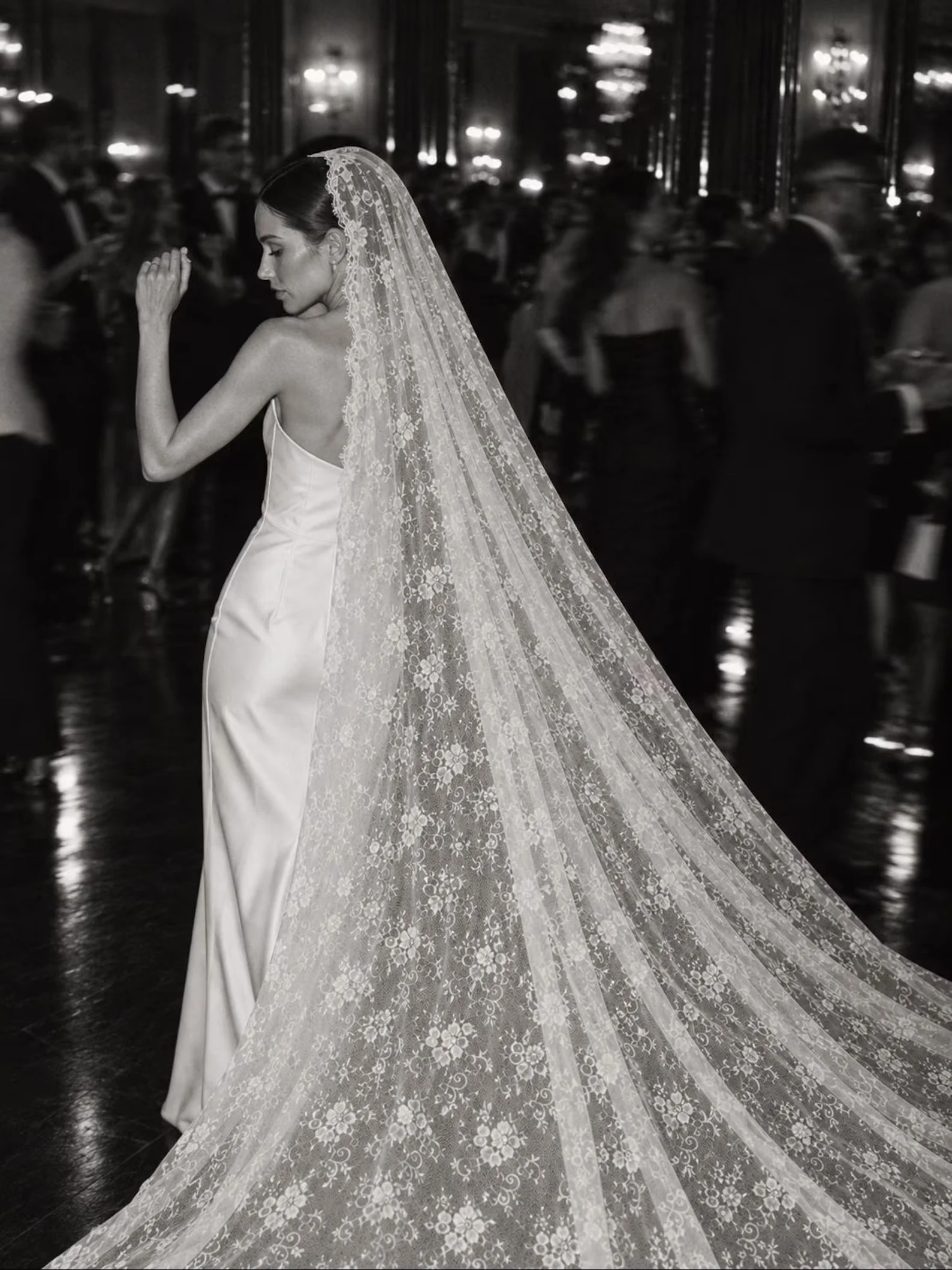 Eine Braut im eleganten Kleid auf einer Feier (S/W). Ihr langer, bodenlaufender Spitzenschleier breitet sich prachtvoll hinter ihr auf dem Tanzboden aus, während sie sich zur Seite dreht.