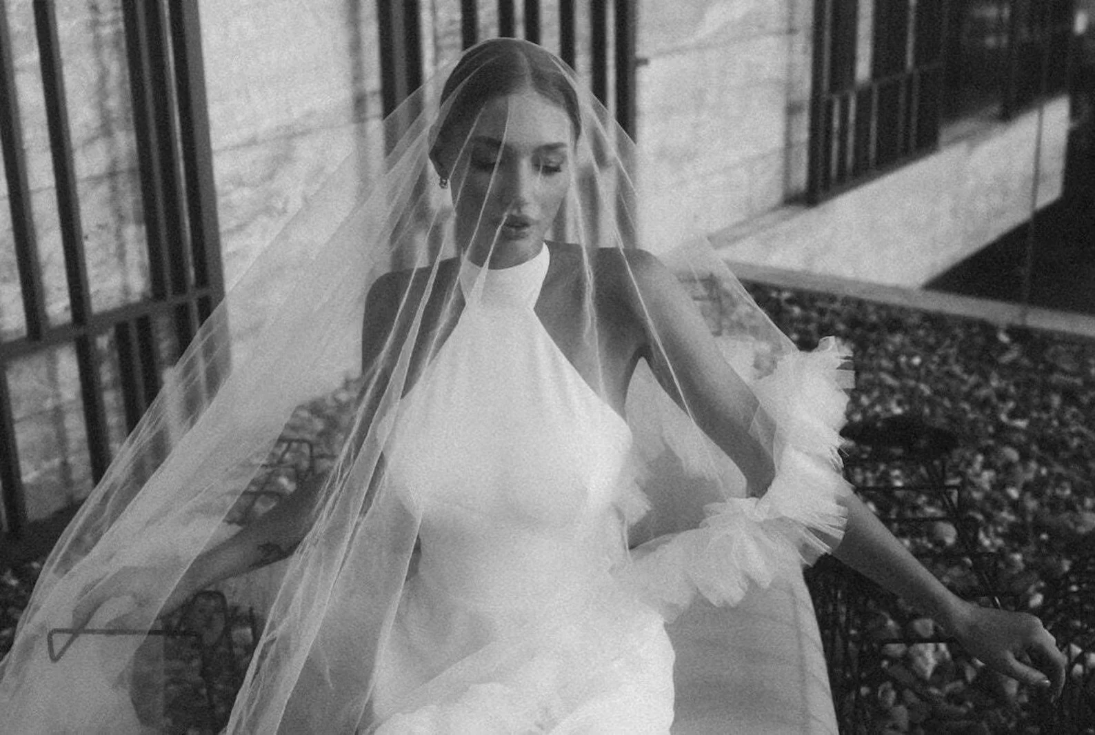 Körniges Schwarz-Weiß-Foto einer Braut in einem hochgeschlossenen Kleid, die einen feinen, transparenten Schleier über Gesicht und Schultern trägt, während sie im Freien sitzt.