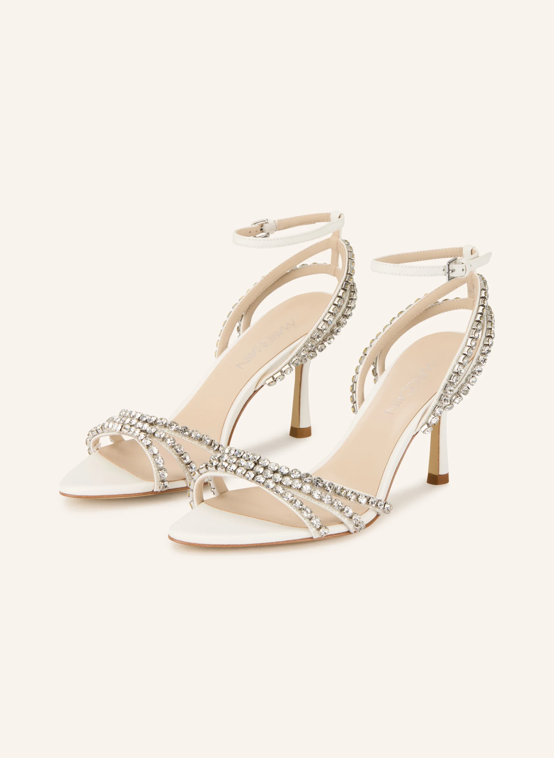 Weiße Sandalen von Marc Cain mit opulentem Kristallbesatz auf den Riemen, Knöchelriemchen und mittelhohem Pfennigabsatz vor hellem Hintergrund.