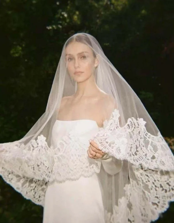 Elegante Braut im trägerlosen Hochzeitskleid mit einem feinen Tüllschleier vor dem Gesicht, der am Saum mit opulenter, breiter Spitze verziert ist. Inspiration für den Brautschleier-Guide.