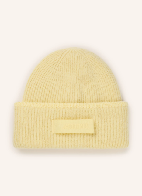 Buttergelbe Strickmütze aus weichem Rippstrick mit breitem Umschlag. Flauschige Kopfbedeckung mit rechteckigem Jacquemus Logo Patch vorne mittig vor hellem Hintergrund.