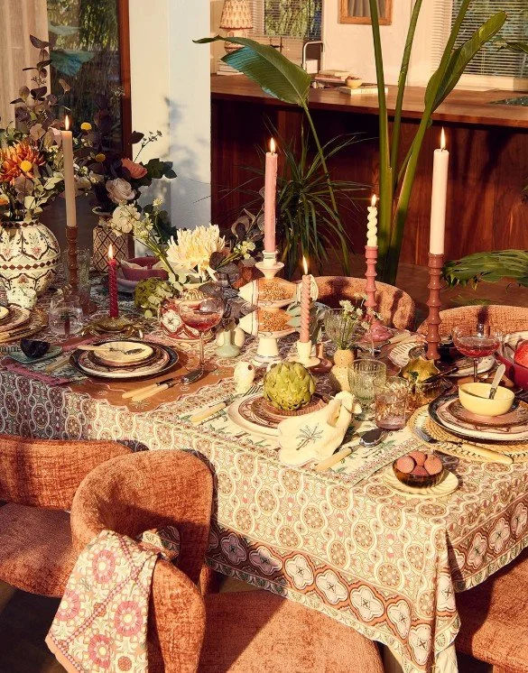 Opulent gedeckte Ostertafel im Sissy-Boy Stil mit gemusterter Tischdecke in warmen Tönen, fischförmigen Kerzenhaltern, gedrehten Kerzen und üppigem Blumenschmuck.