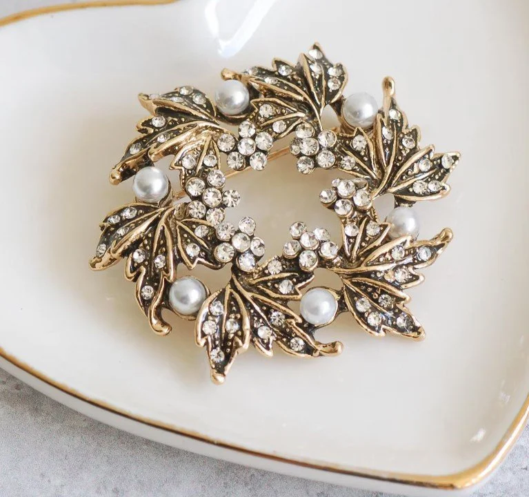 Eine kreisförmige, goldfarbene Brosche in Form von Ahornblättern, die mit kleinen weißen Perlen und glitzernden Kristallen besetzt ist. Das Schmuckstück liegt in einer weißen, herzförmigen Schale mit Goldrand. Hochwertiger Brosche Vintage Schmuck.