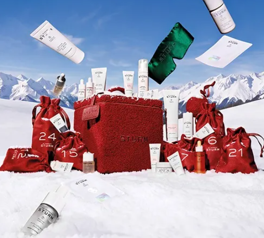 Ein Dr. Barbara Sturm Adventskalender vor verschneiter Bergkulisse. Eine flauschige rote Box und nummerierte Stoffsäckchen werden von verschiedenen Hautpflegeprodukten im Schnee und in der Luft umrahmt.