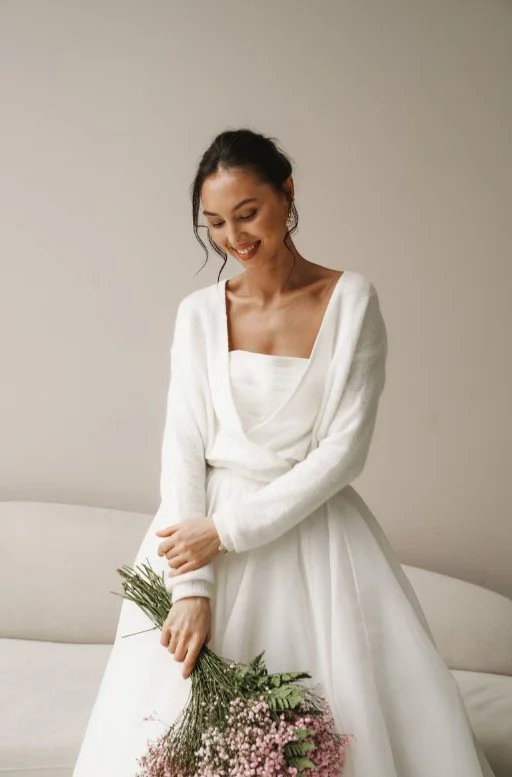 Braut mit lockerem, weißem Wickelpullover über einem trägerlosen Kleid hält einen Strauß aus rosa Schleierkraut, lächelnd vor hellem Hintergrund.