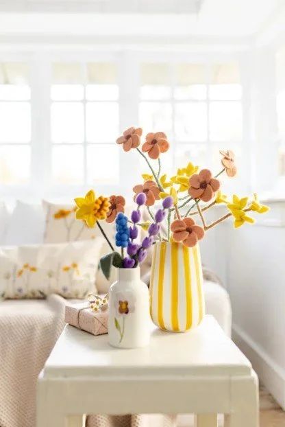 Helle Osterdekoration: Eine gelb-weiß gestreifte Vase mit bunten Filzblumen (Narzissen und Vergissmeinnicht) auf einem weißen Beistelltisch im Gegenlicht.
