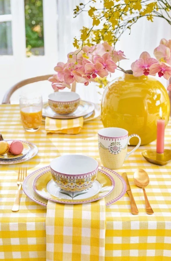 Festlich gedeckter Ostertisch in Gelb: Karierte Tischdecke, eine gelbe Vase mit rosa Orchideen und buntes, florales Geschirr von Pip Studio.