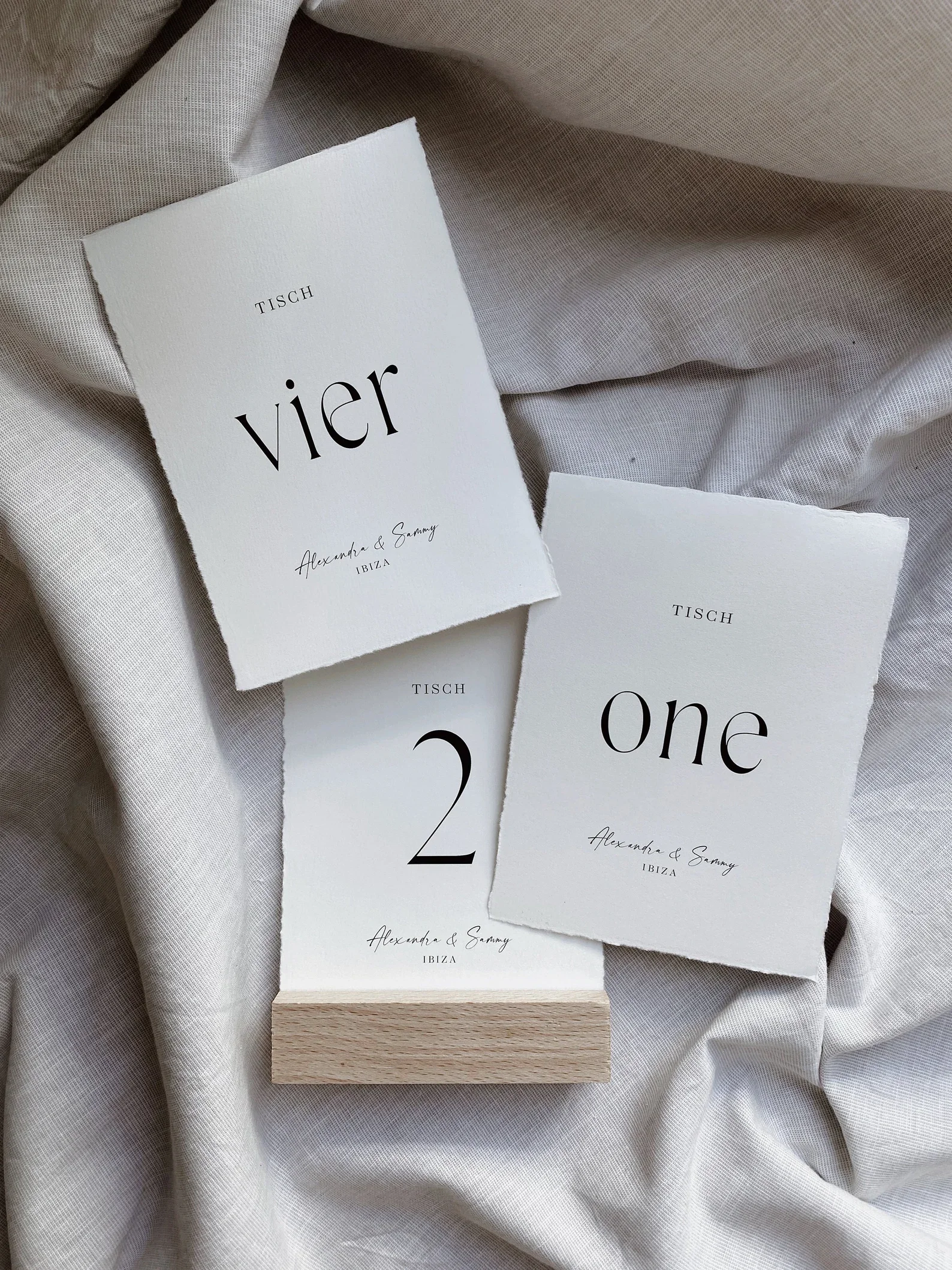 Drei minimalistische Hochzeitskarten aus weißem Büttenpapier mit modernem schwarzem Aufdruck („vier“, „one“, „2“) in einem schlichten Holzaufsteller.