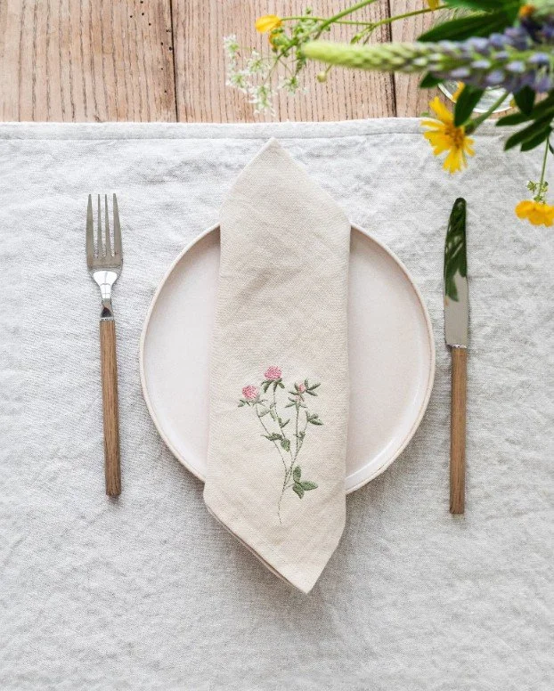 Minimalistisches Tischgedeck für den Frühling: Eine beige Leinenserviette mit bestickten rosa Kleeblüten liegt auf einem hellen Teller, flankiert von Besteck mit Holzgriffen auf einer grauen Leinentischdecke.