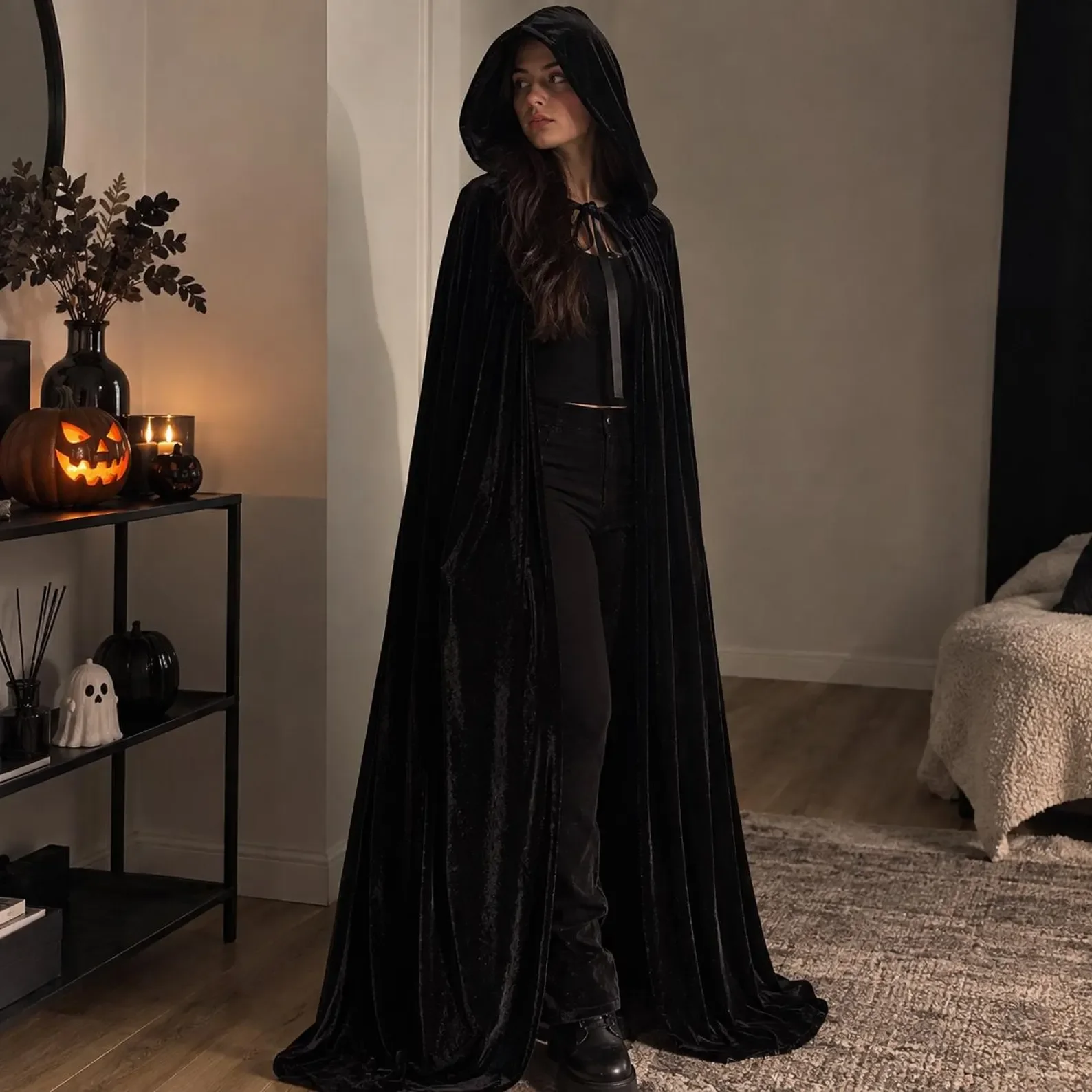 Eine Frau in einem bodenlangen, schwarzen Samtumhang mit aufgesetzter Kapuze. Sie steht in einem Wohnraum, der im Hintergrund mit Halloween-Elementen wie einem leuchtenden Kürbis dekoriert ist.