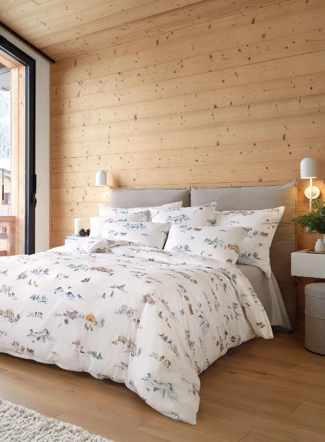 Helles Schlafzimmer mit Holzverkleidung und einem Bett, bezogen mit weißer Bettwäsche mit winterlichen Berg- und Dorfmotiven, vielen Kissen und gepolstertem Kopfteil.