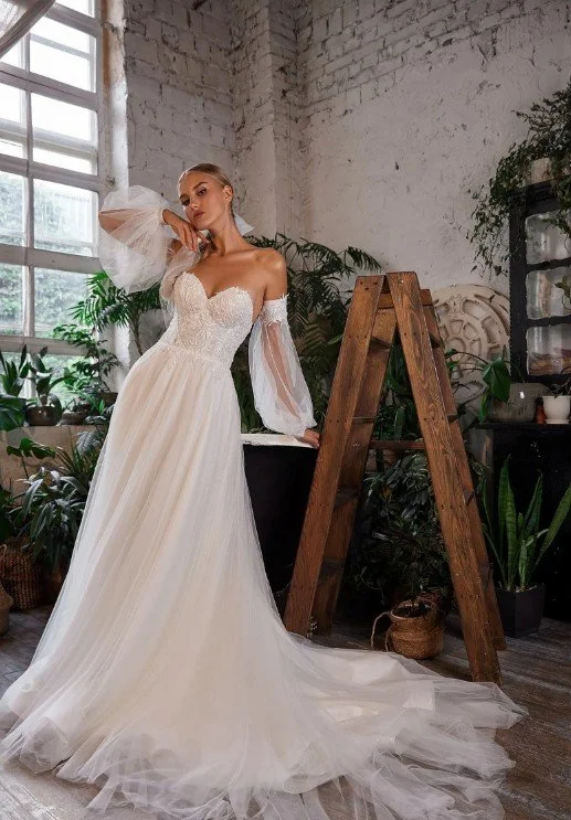 Frau in einem schulterfreien weißen Brautkleid mit transparenten Ärmeln und Tüllrock, stehend in einem rustikalen Raum mit Pflanzen und Holzleiter.