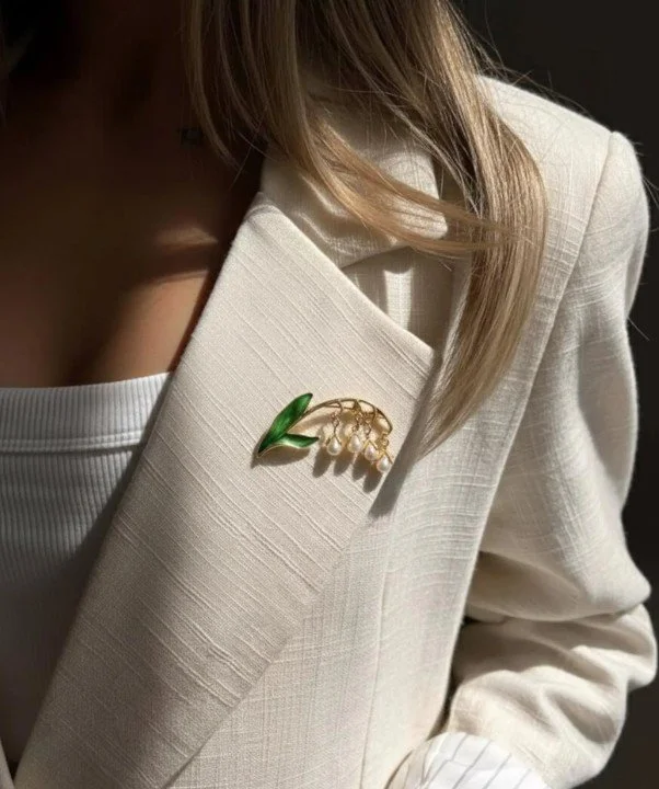 Nahaufnahme einer Frau in einem cremefarbenen Leinenblazer. Am Revers ist eine goldfarbene Brosche in Form eines Maiglöckchens mit grünen Blättern und kleinen weißen Perlen befestigt. Sanftes Sonnenlicht fällt auf den Stoff und den Schmuck.