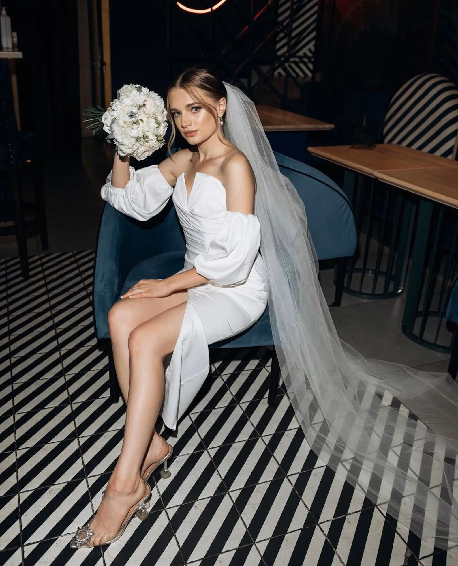 Eine sitzende Braut in einem schulterfreien Kleid mit Beinschlitz und langem Schleier hält einen Strauß weißer Blumen, während sie auf einem gemusterten Schwarz-Weiß-Boden posiert.