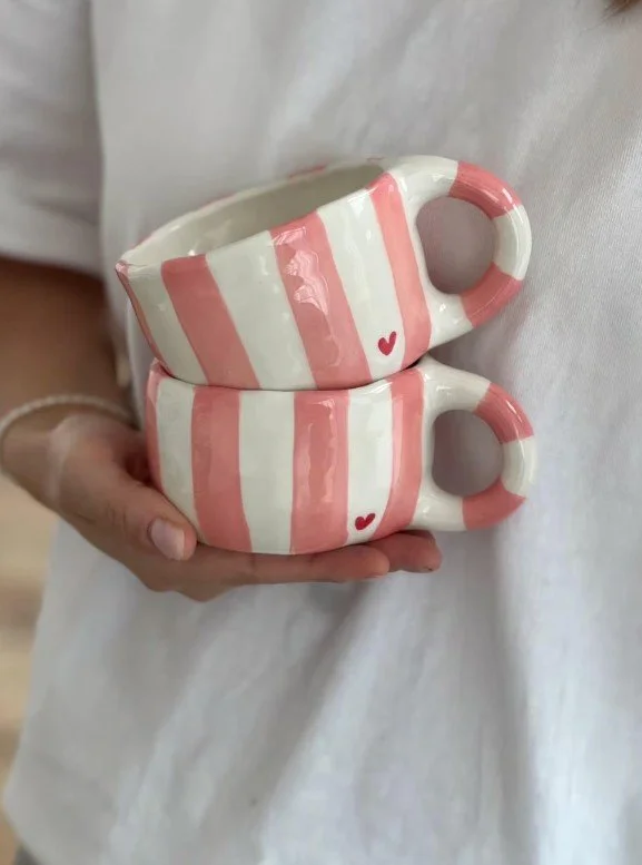 Zwei handgefertigte rosa-weiß gestreifte Tassen mit kleinem roten Herz, übereinander gestapelt und von einer Person in einem weißen Shirt gehalten.
