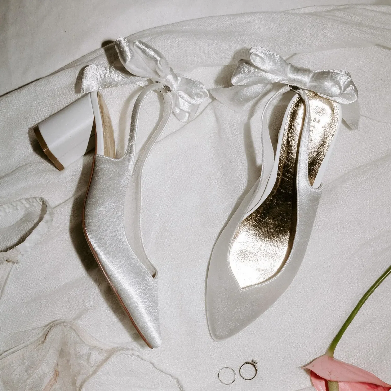 Zwei spitze Satin-Slingback-Pumps mit Samt-Schleifen am Knöchel und goldener Innensohle liegen auf weißem Leinenstoff, daneben Eheringe und eine rosa Blüte.