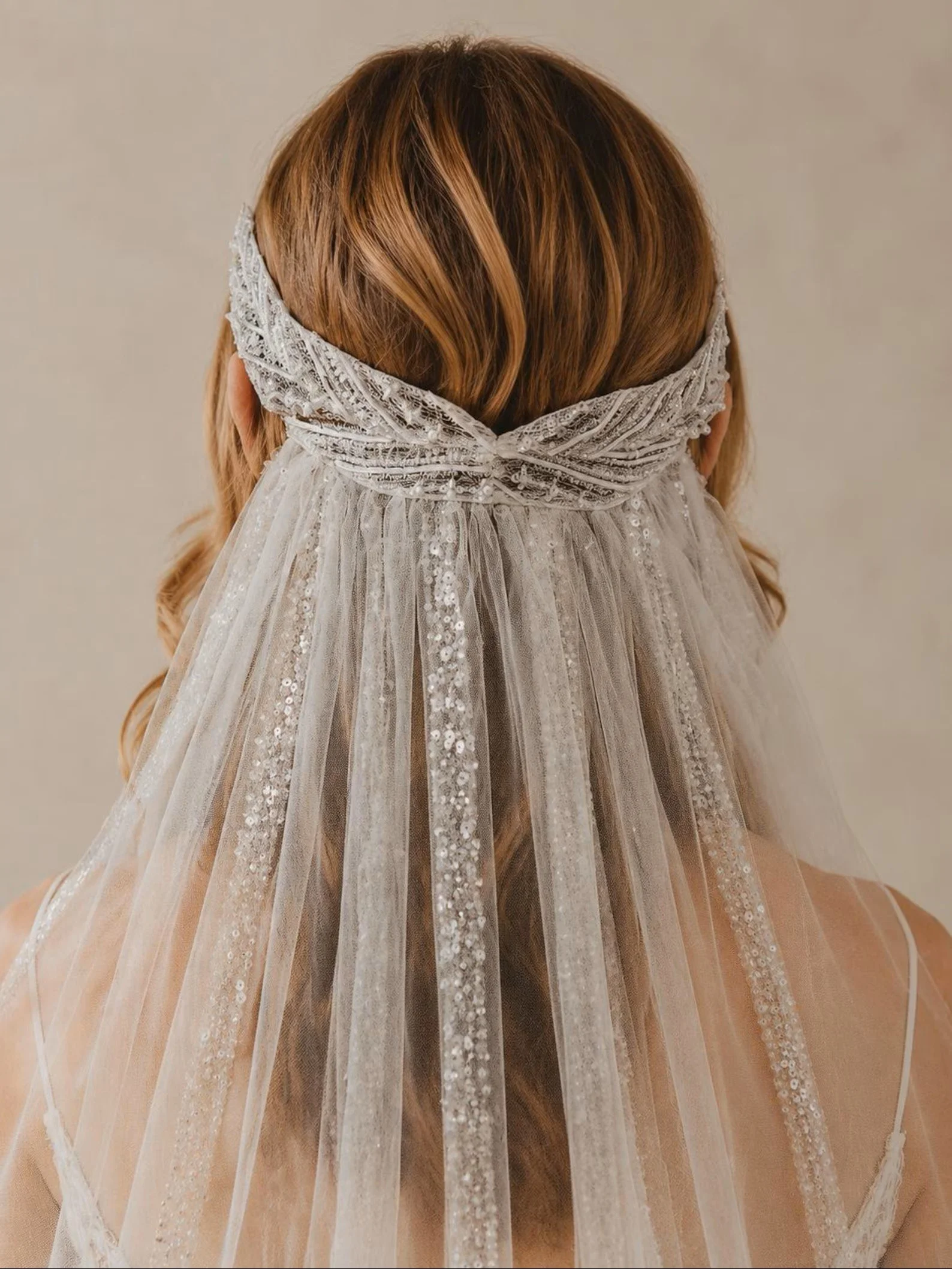Rückansicht einer Braut mit rötlichem Haar. Sie trägt ein verziertes, silbernes Stirnband am Hinterkopf, von dem ein feiner Tüllschleier mit glitzernden Pailletten-Streifen herabfällt.