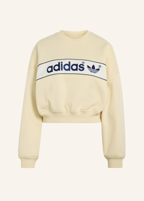 Cremeweißer cropped Adidas Pullover mit Rundhalsausschnitt. Dunkelblauer Adidas Schriftzug und Trefoil Logo auf weißem Bruststreifen. Rippbündchen an Hals Ärmeln und Saum.