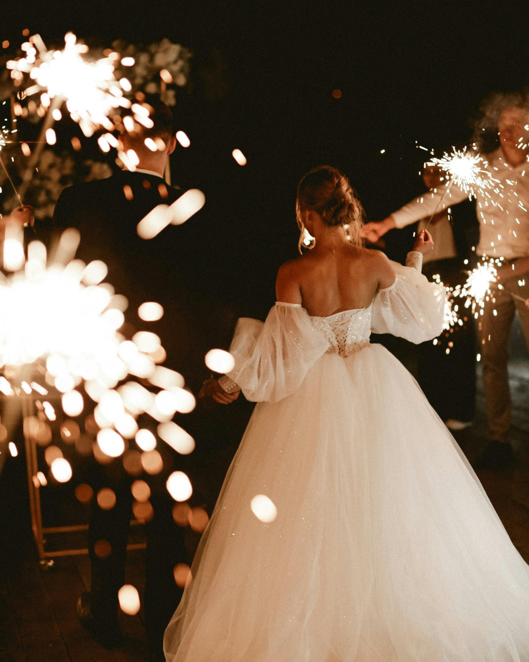 Braut und Bräutigam von hinten bei einer Adults-only-Hochzeit am Abend, umgeben von Gästen mit Wunderkerzen. Die Braut trägt ein schulterfreies Hochzeitskleid mit opulenten Tüllärmeln.