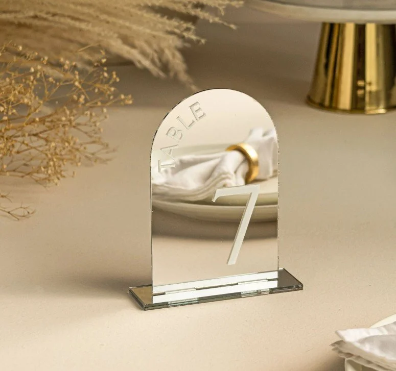 Eine verspiegelte Tischnummer in Bogenform mit der Gravur „TABLE 7“, die auf einem beigen Untergrund steht und die Dekoration reflektiert.