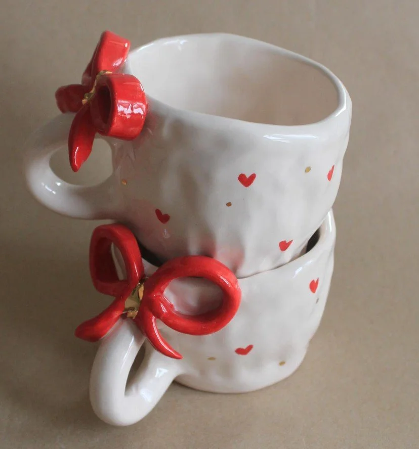 Zwei übereinander gestapelte weiße Keramiktassen mit roten Herz-Mustern und einer markanten roten Schleife am Henkel als Geschenkidee.