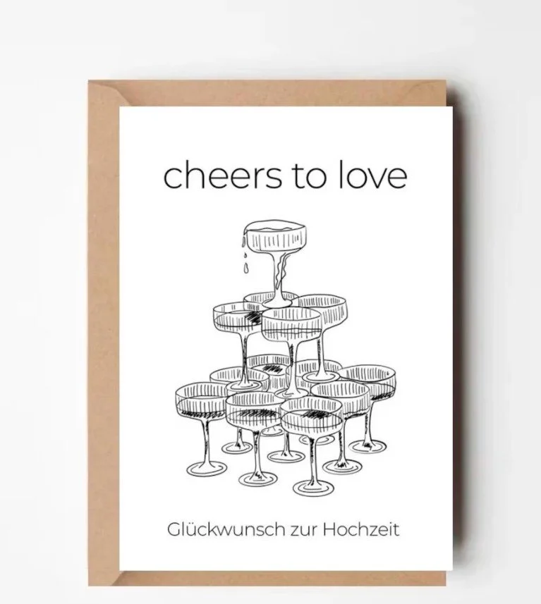 Minimalistische Hochzeitskarte mit einer schwarz-weißen Zeichnung einer Champagner-Pyramide und dem Text „cheers to love“ sowie „Glückwunsch zur Hochzeit“ auf einem braunen Umschlag.