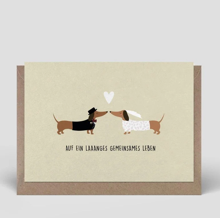 Lustige Glückwunschkarte zur Hochzeit in Beige mit zwei Dackeln, die als Braut und Bräutigam verkleidet sind. Über den Hunden schwebt ein Herz und darunter steht das Wortspiel: „AUF EIN LAAANGES GEMEINSAMES LEBEN“.