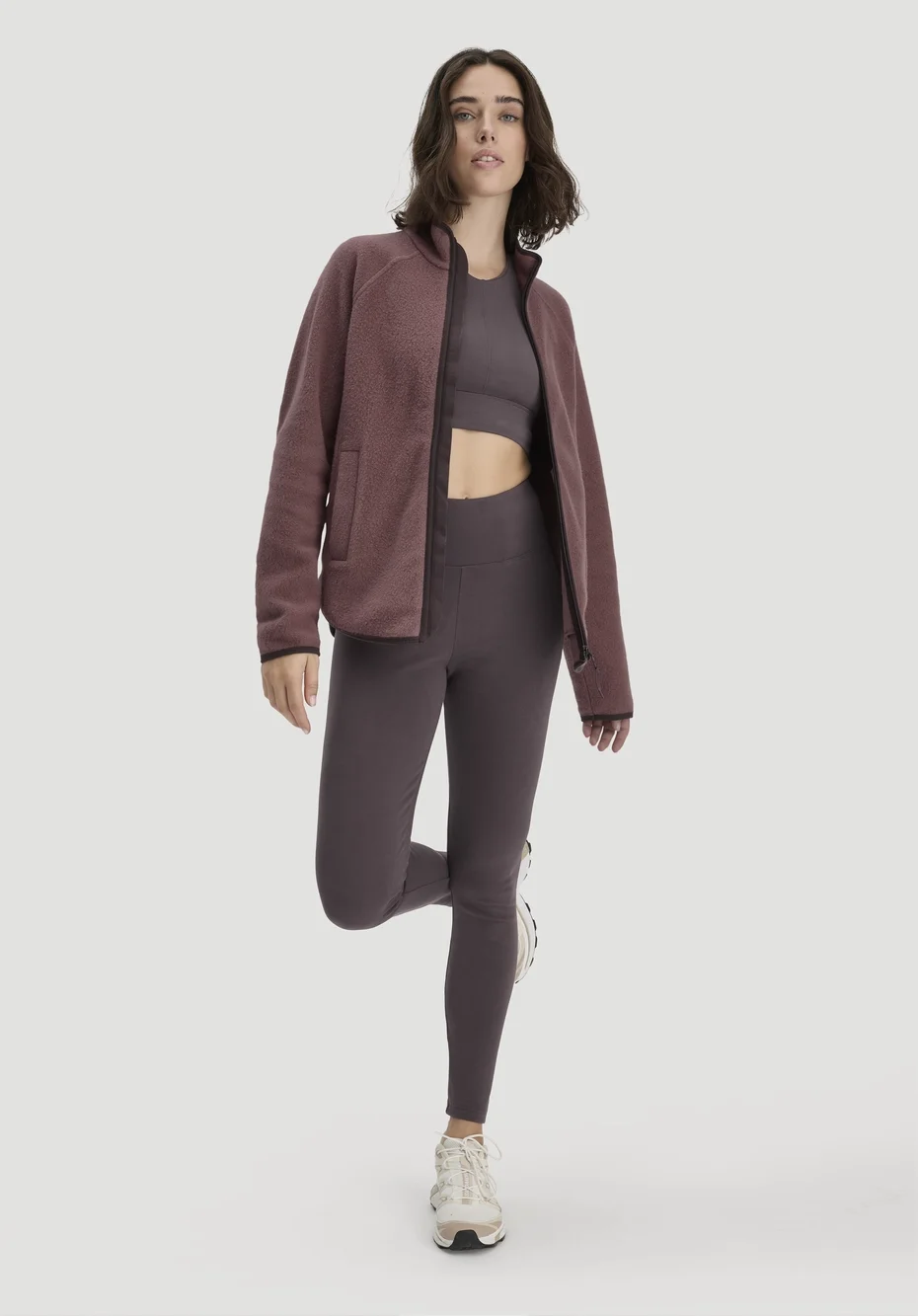Frau in weinroter Fleecejacke über dunkelviolettem Sport-Top und Leggings mit hellen Sneakern vor hellem Hintergrund