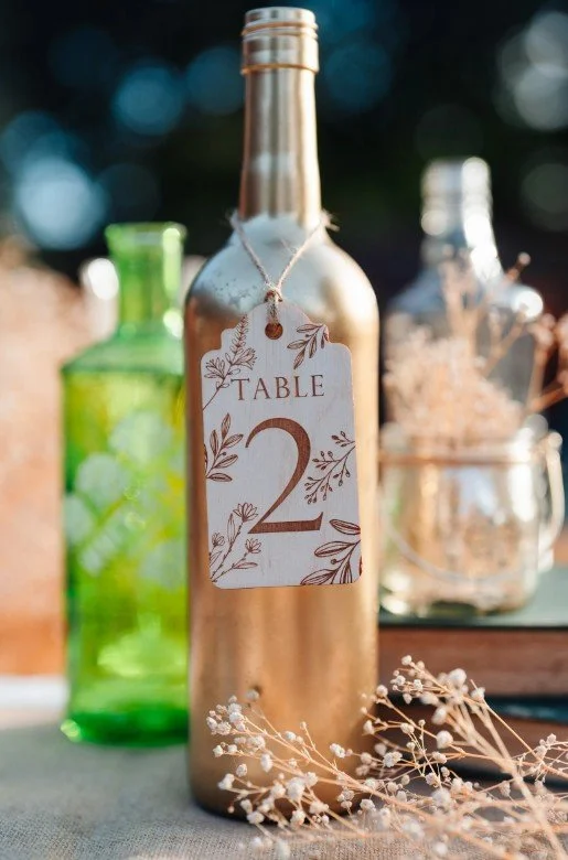 Ein hölzerner Geschenkanhänger mit der Aufschrift „TABLE 2“ und floralen Mustern, der mit einer Schnur um den Hals einer goldfarbenen Weinflasche gebunden ist.