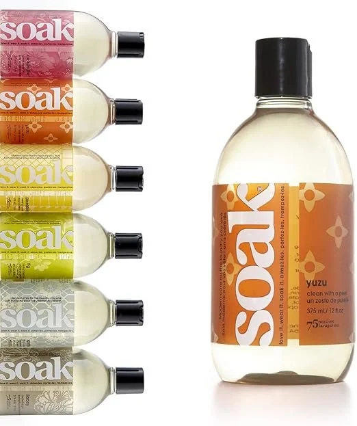 Rechts steht eine große, transparente Flasche des Feinwaschmittels „soak“ in der Duftrichtung Yuzu. Links daneben zeigen sechs übereinandergestapelte, bunte Flaschen die weiteren Duftvarianten der Marke.