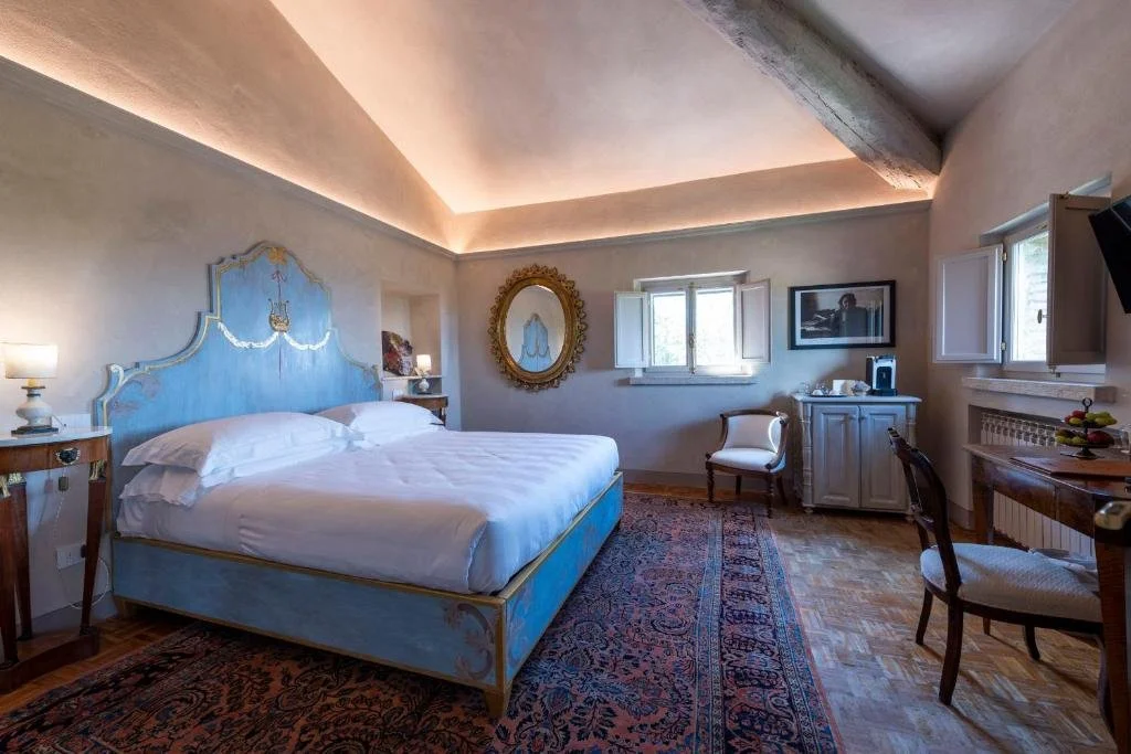 Foto eines eleganten Schlafzimmers mit schräger Decke, einem rustikalen Holzbalken und indirekter Beleuchtung. Im Zentrum steht ein großes Bett mit einem kunstvoll hellblau-gold verzierten Rahmen auf einem großen Orientteppich über Holzparkett.