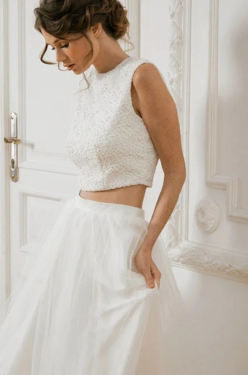 Zweiteiliges Brautkleid (Two-Piece) mit einem hochgeschlossenen, ärmellosen Spitzen-Crop-Top und einem langen, weichen Tüllrock vor einer weißen Vintage-Tür.