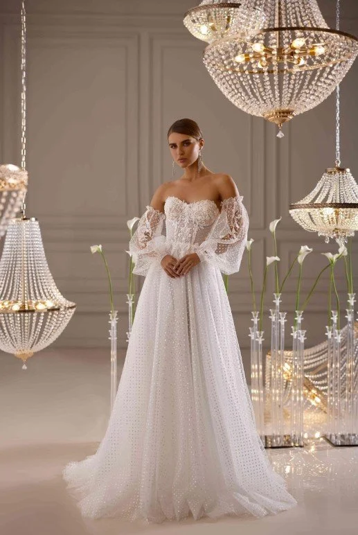 Frau in einem schulterfreien weißen Brautkleid mit transparenten Puffärmeln, stehend in einem eleganten Raum mit Kristallkronleuchtern und Blumen.