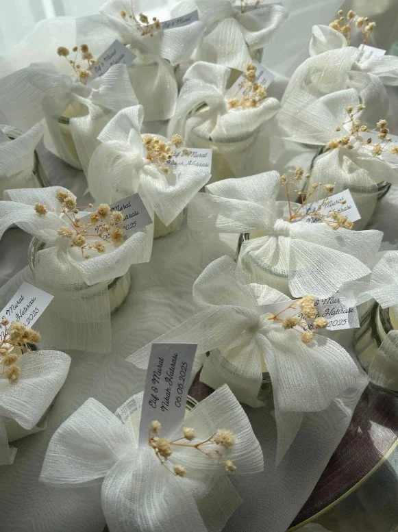 Kleine Hochzeitskerzen als Gastgeschenk in Gläsern, elegant verpackt mit weißer Schleife, Trockenblumen und Namensetikett.